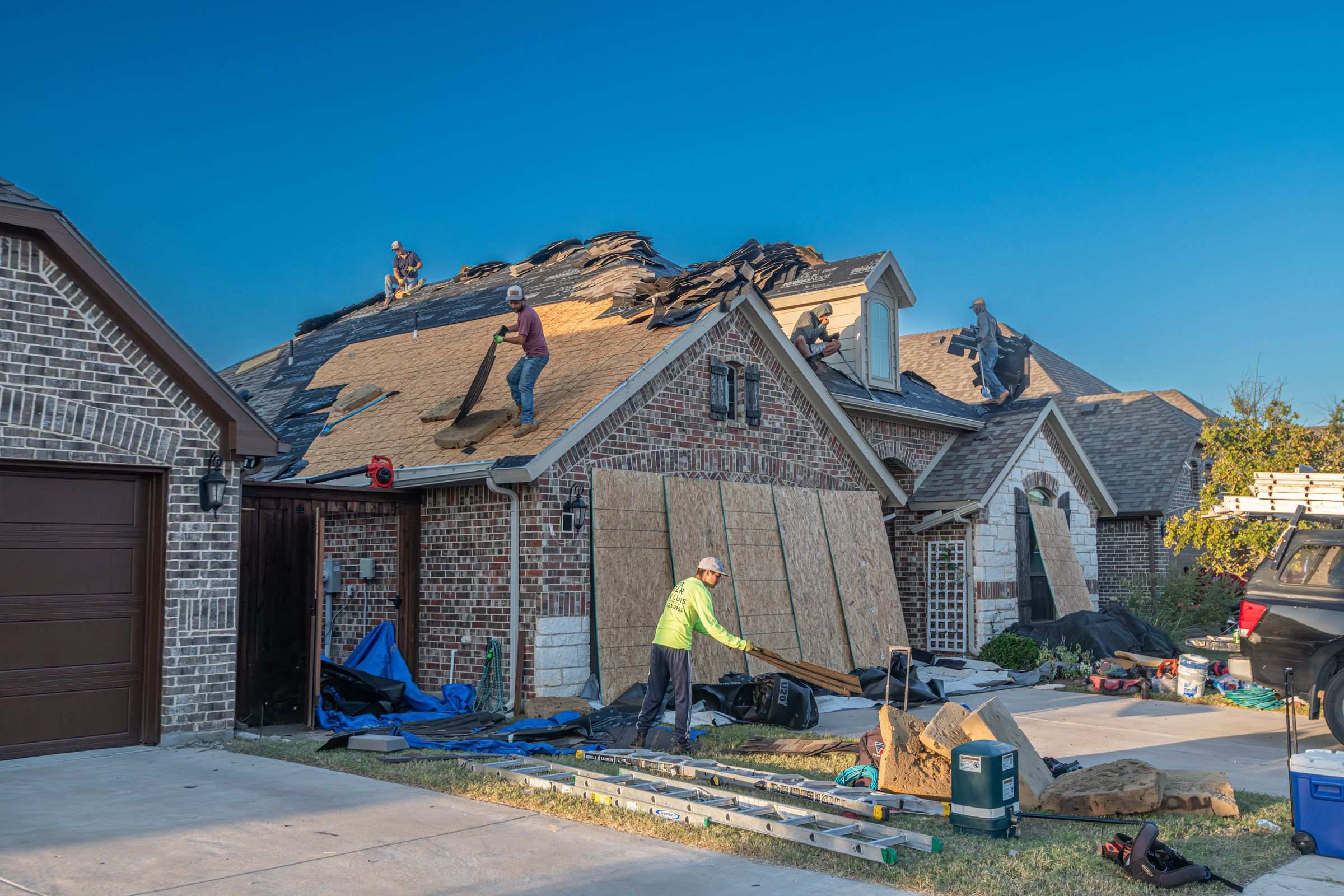 Kestrel crew tearing off an old roof on a brick home; new plywood decking leans against the garage